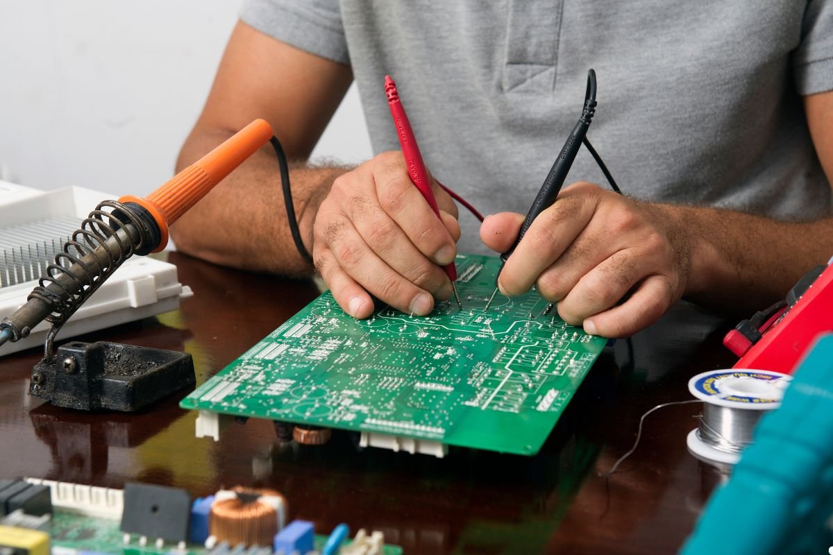 A man carefully works on a circuit board, using a pair of scissors to trim components or wires.